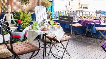 A patio with a table set for dining, surrounded by chairs and cushions, with plants and a decorative fence in the background.