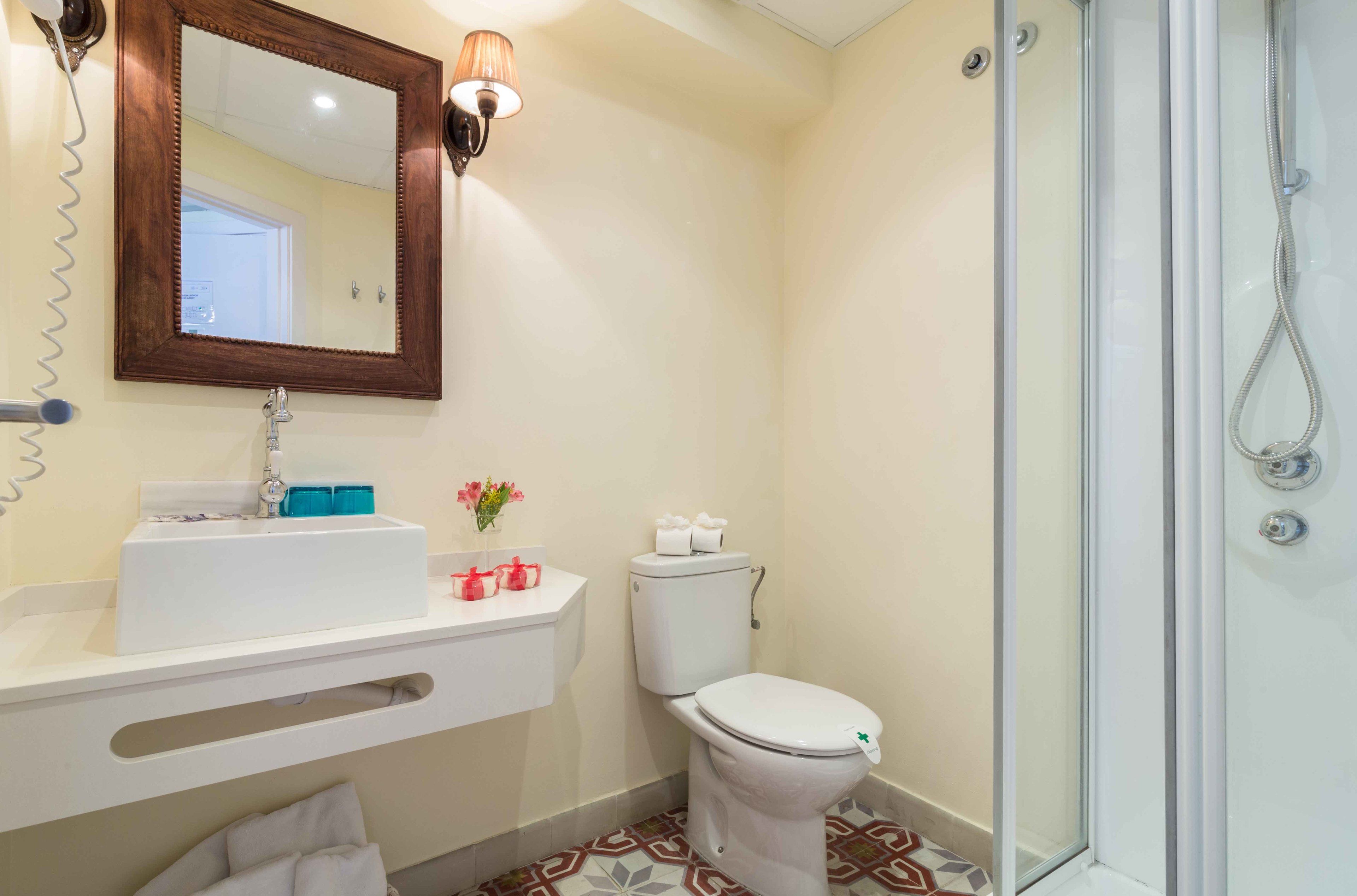 Un baño con lavamanos blanco, espejo y retrete, ducha con puerta de cristal, y piso con diseño.