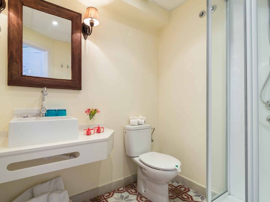 Un baño con lavamanos blanco, espejo y retrete, ducha con puerta de cristal, y piso con diseño.
