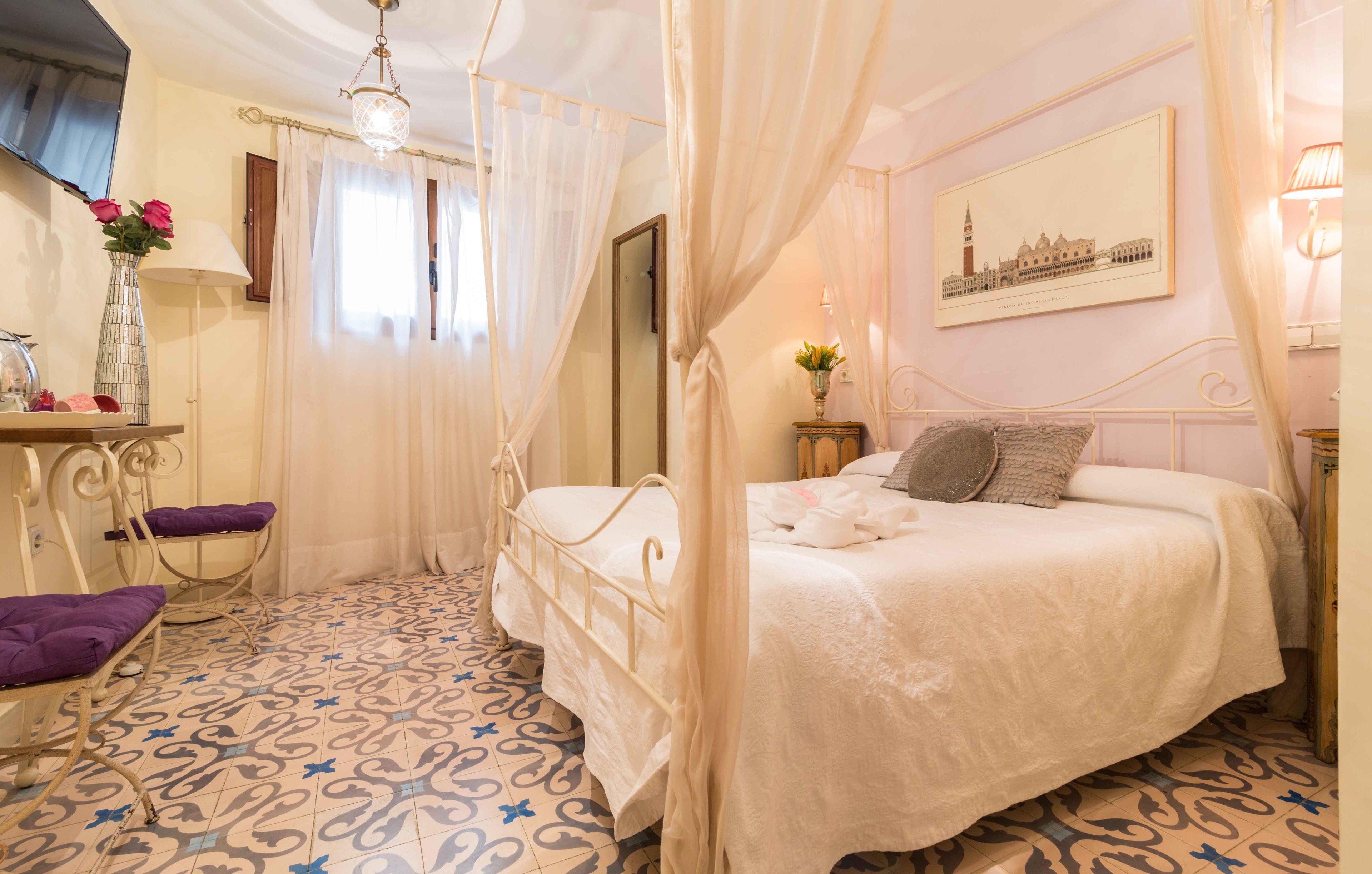 A bedroom with a four-poster bed, a bedside table with a lamp, a chair with a cushion, and a wall-mounted TV.