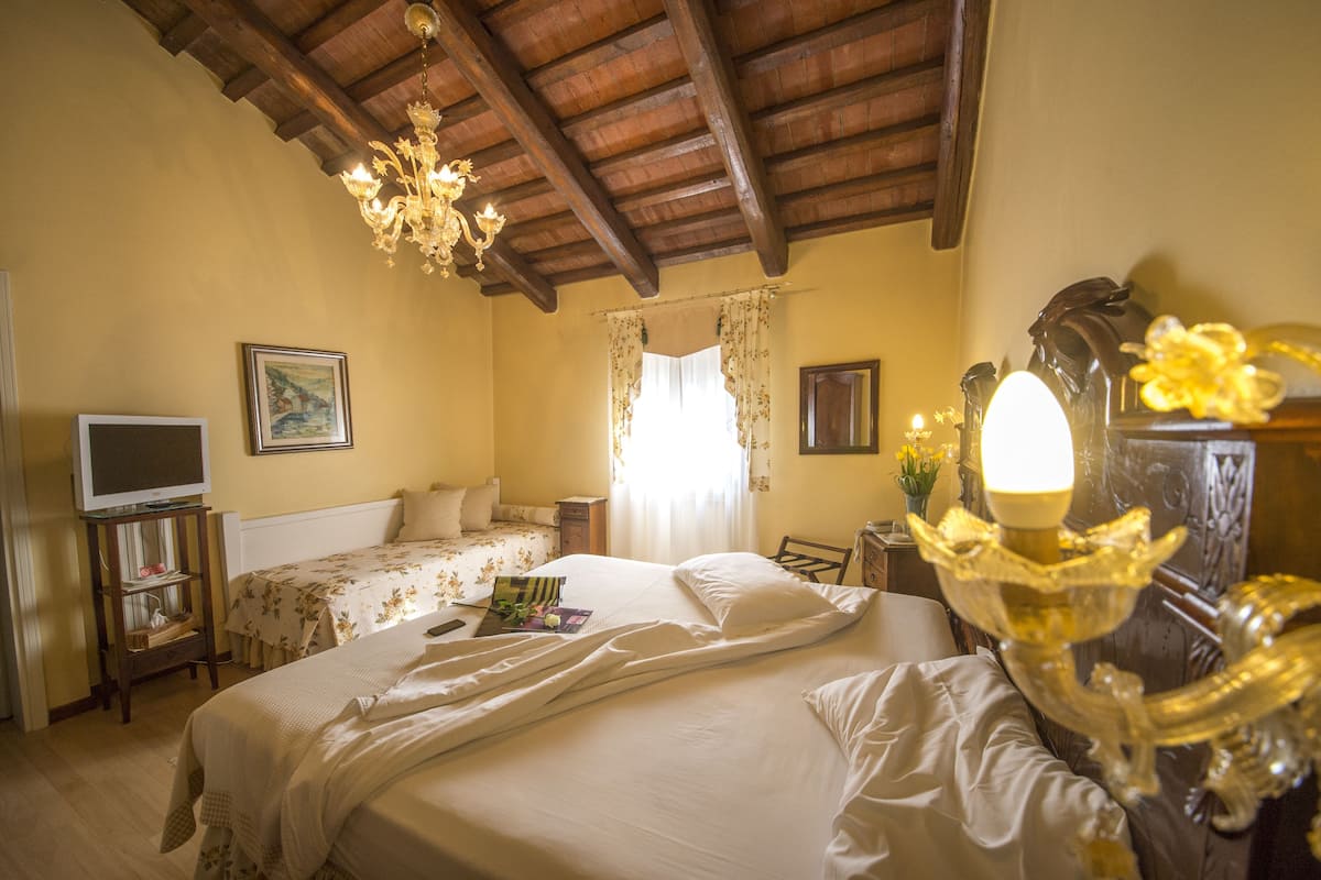 A bedroom with a wooden ceiling, a chandelier, a bed with floral bedding, a television on a stand, and a dresser with a mirror.