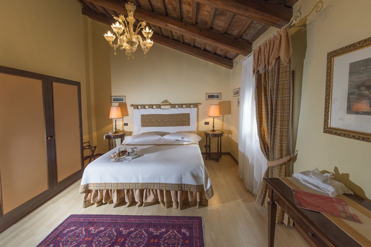 A bedroom with a large bed, two bedside tables with lamps, a chandelier, a wooden ceiling, a plaid curtain, and a framed picture on the wall.