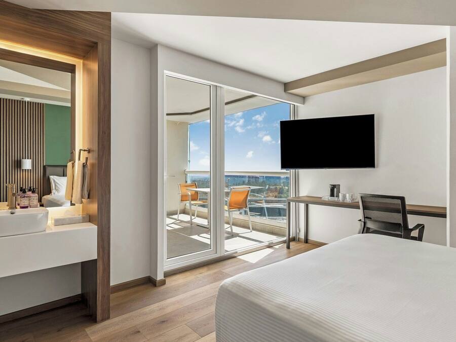 Una camera d'albergo con un letto grande, una TV a muro, un balcone con vista e un bagno con mobile e specchio.