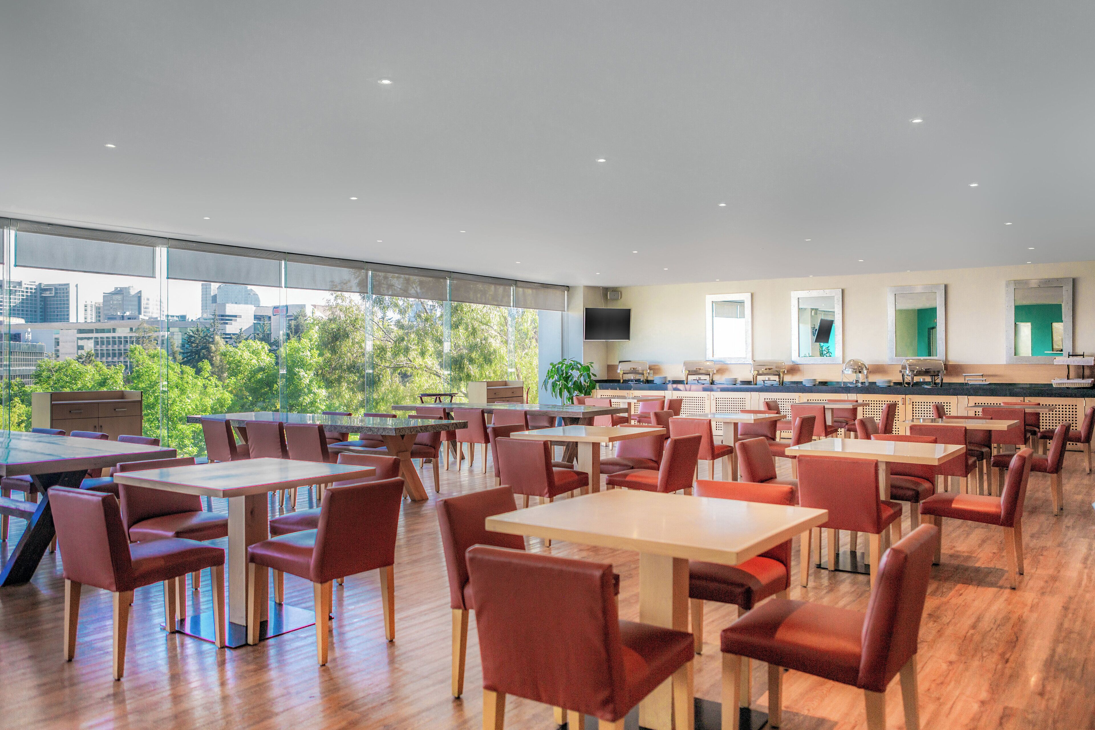 A modern dining area with multiple tables and chairs, large windows offering a city view, and a kitchen area in the background.