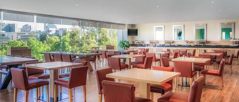 A modern dining area with multiple tables and chairs, large windows offering a city view, and a kitchen area in the background.