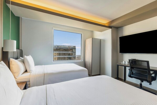 A hotel room with two beds, a desk with a chair, a TV, and a window with a view of buildings.