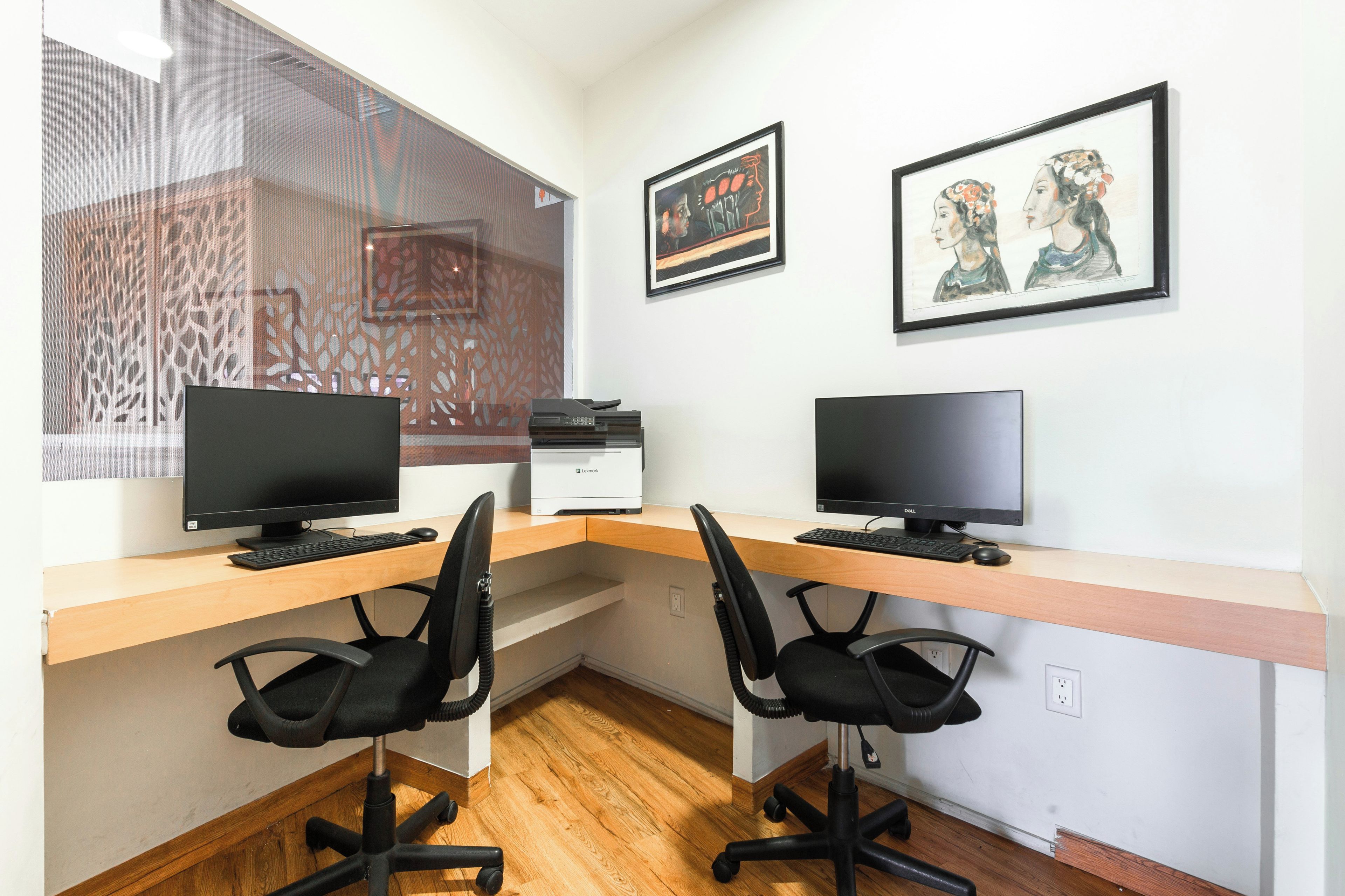 A workspace with two desks, each with a computer monitor, keyboard, and chair. There are two framed artworks on the wall.