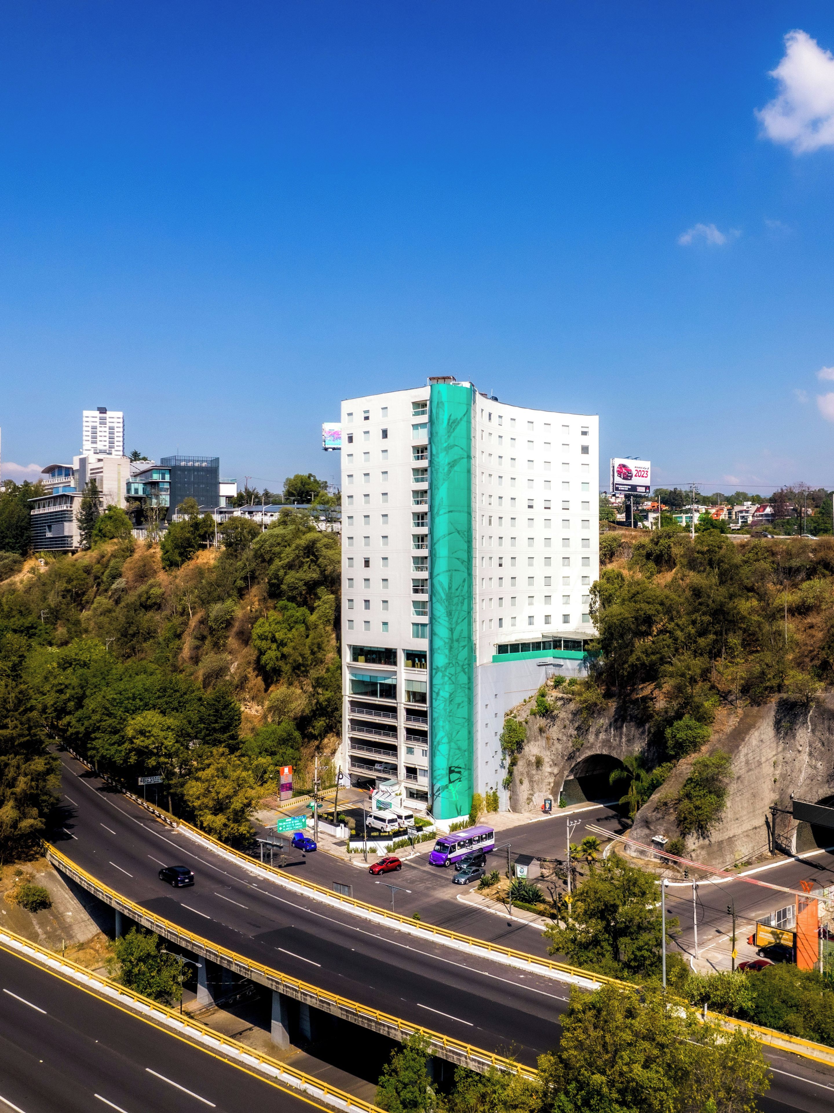 Un edifici de diversos pisos amb un element vertical verd destacat, situat a prop d'una autopista on es veuen vehicles.