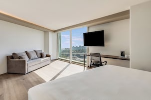 Une chambre d’hôtel moderne dotée d’un grand lit, d’un canapé, d’un bureau avec une chaise, d’une télévision à écran plat et d’une grande fenêtre donnant sur la ville.