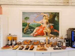 Ein Frühstücksbuffet mit Gebäck, Brot und Aufstrichen, darüber ein klassisches Gemälde.