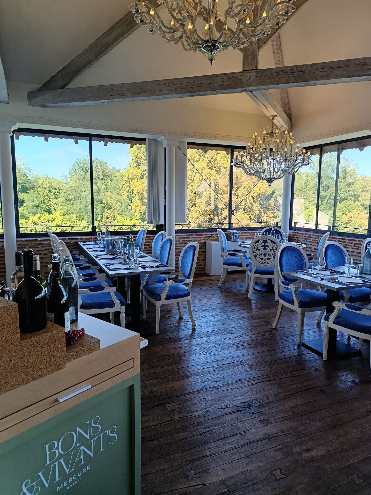 Restaurante con techo alto, candelabros y grandes ventanales que ofrecen vista a árboles. Las mesas están dispuestas con sillas azules y copas de vino.