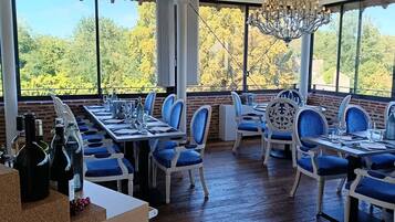 Restaurante con techo alto, candelabros y grandes ventanales que ofrecen vista a árboles. Las mesas están dispuestas con sillas azules y copas de vino.