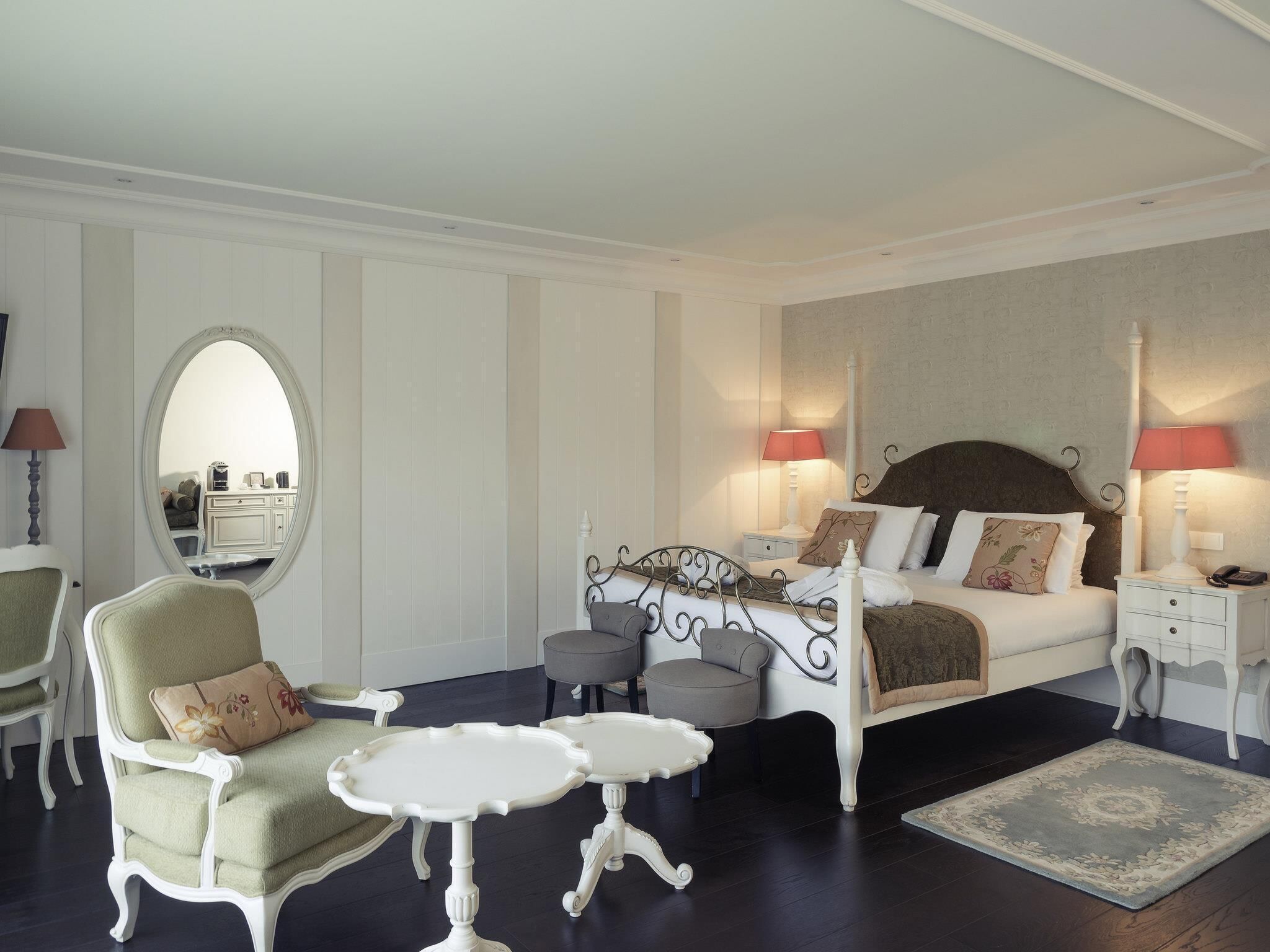 Ein Schlafzimmer mit einem Bett, zwei Nachttischen, einem Sessel, einem Spiegel und einem kleinen Tisch.