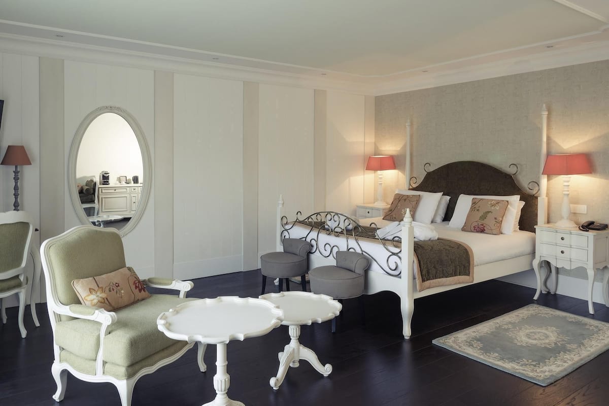 Ein Schlafzimmer mit einem Bett, zwei Nachttischen, einem Sessel, einem Spiegel und einem kleinen Tisch.