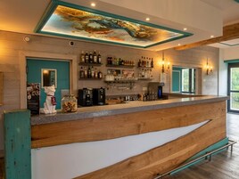 Una zona de bar con mostrador de madera, estantes con botellas, una máquina de café y un mural en el techo.