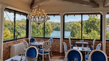 Un'area pranzo con tavoli e sedie, ampie finestre vista su un lago e una foresta, e un lampadario.