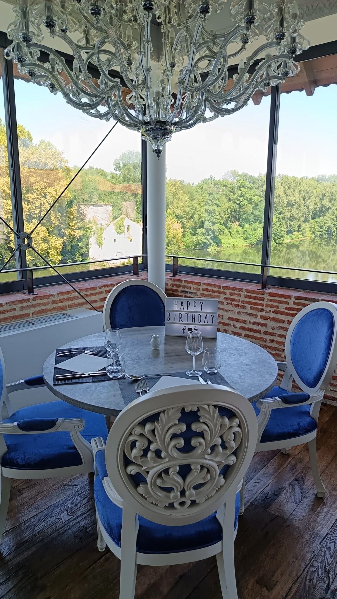 A dining area with a round table, blue upholstered chairs, a chandelier, and a view of greenery through large windows.