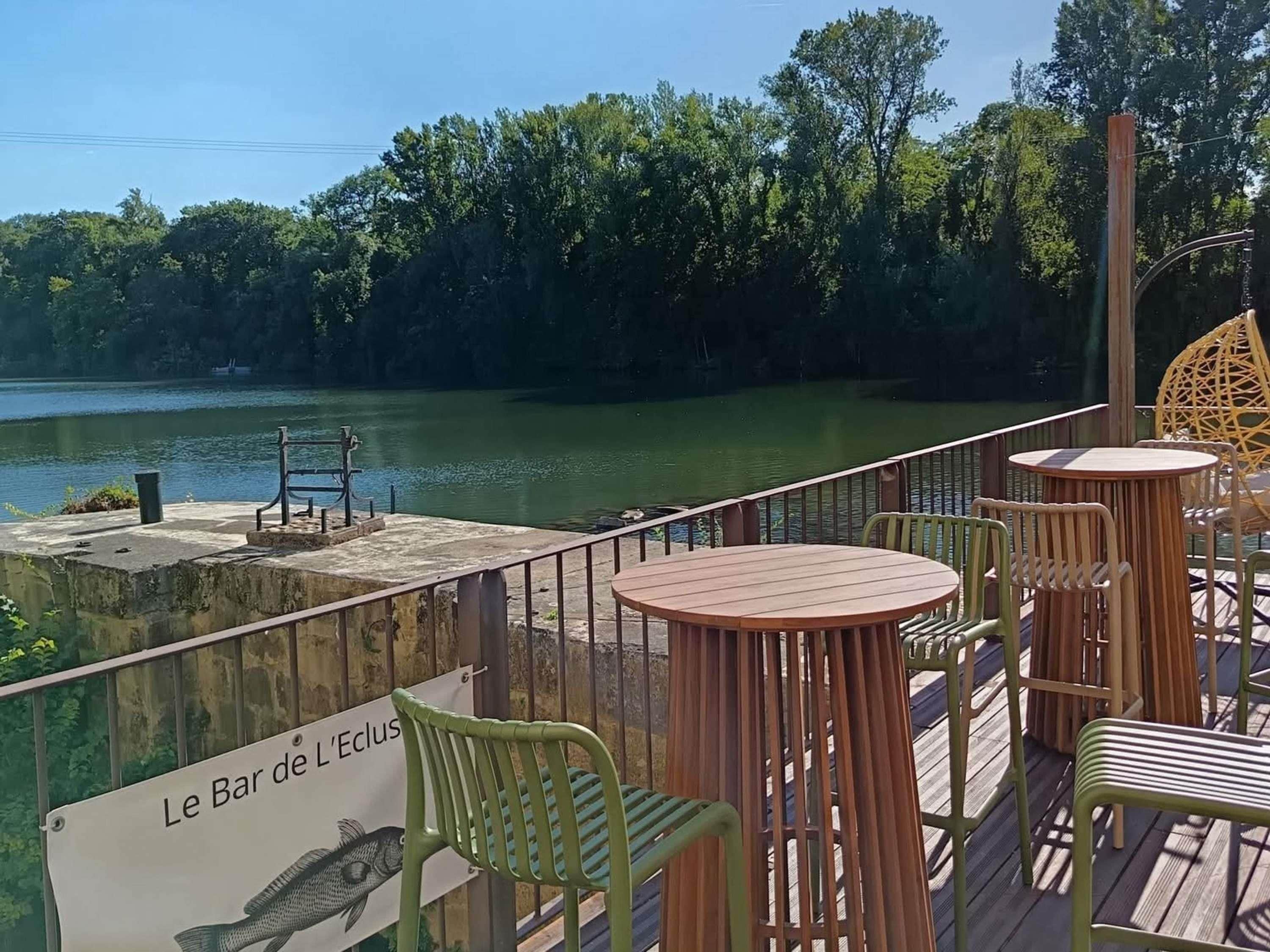 Una terraza de madera con mesas y sillas da a un cuerpo de agua tranquilo, con un letrero que dice 'Le Bar de L'Eclus'.