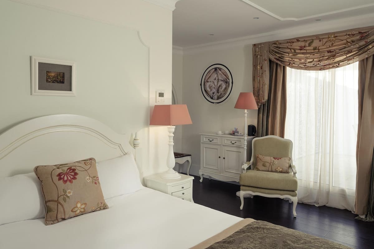 Ein Schlafzimmer mit Bett, Nachttischen, einem Sessel, einem Schminktisch und einem Fenster mit Vorhängen.