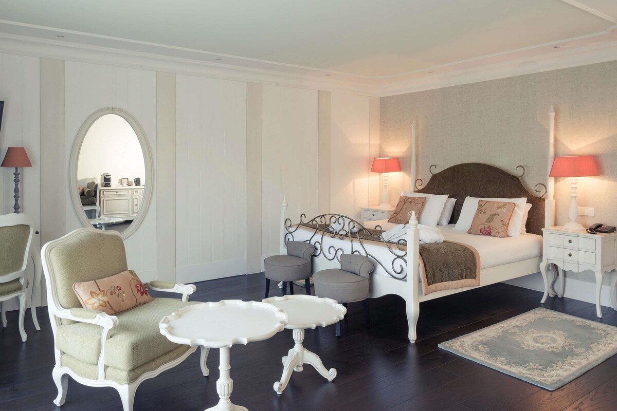 A bedroom with a four-poster bed, two bedside tables, a chair, a small round table, and a mirror.
