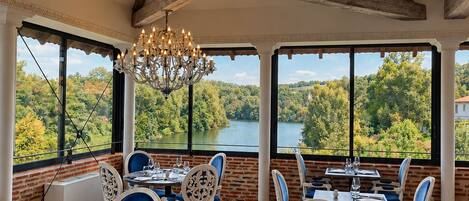 A dining area with tables and chairs, large windows with a view of a lake and trees, and a chandelier.