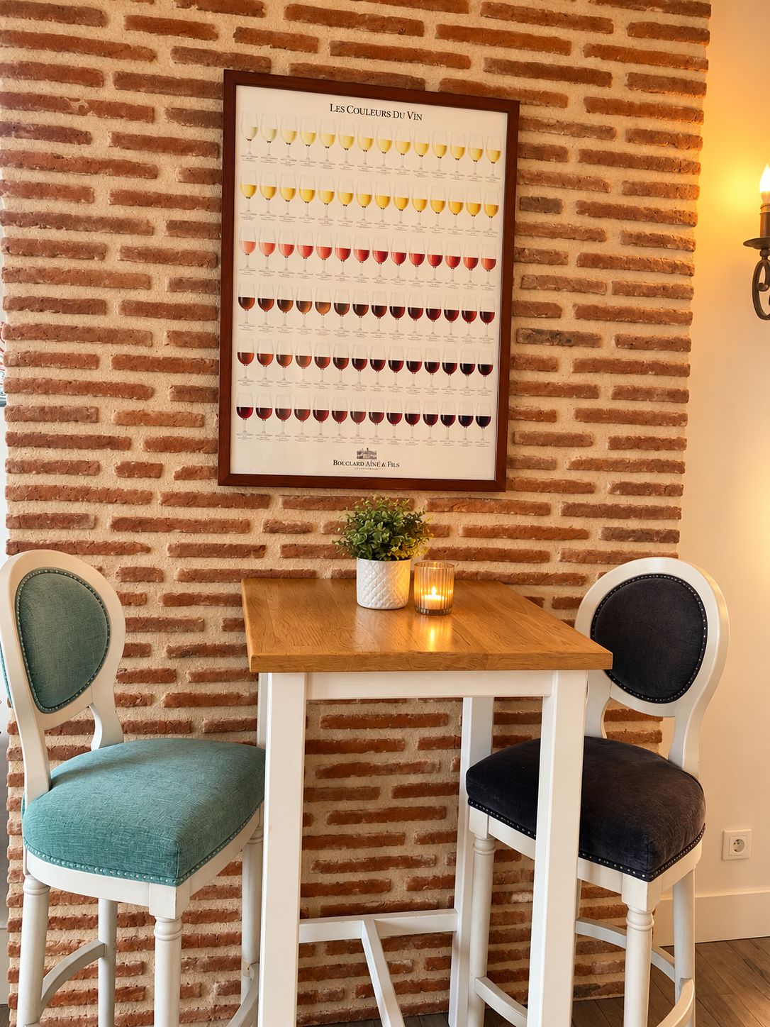 A brick wall with a framed poster of wine colors, a wooden table, and two chairs.