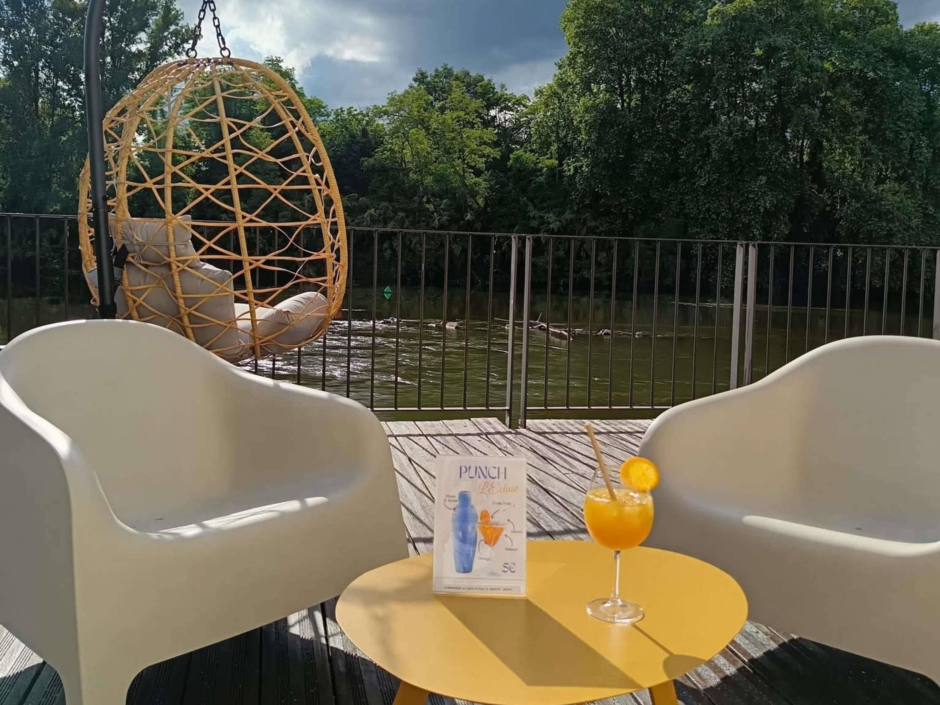 Een terras met twee witte stoelen, een hangstoel, een tafel met een drankje en een menu, en uitzicht op een waterpartij met bomen op de achtergrond.
