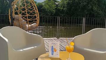 Een terras met twee witte stoelen, een hangstoel, een tafel met een drankje en een menu, en uitzicht op een waterpartij met bomen op de achtergrond.