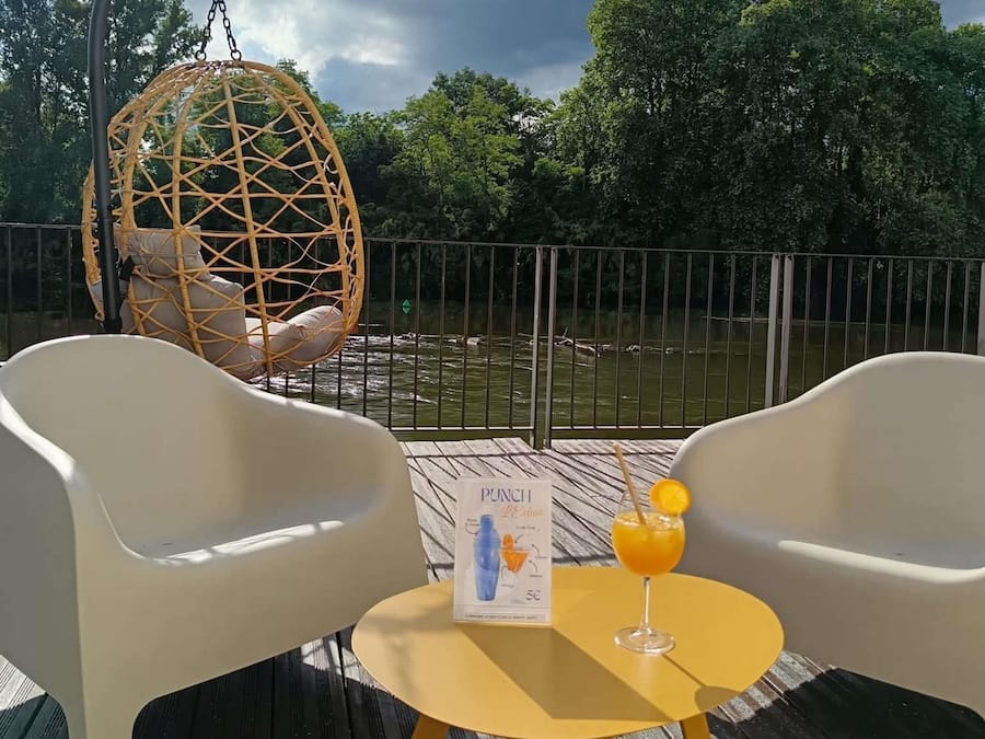 Una terraza con dos sillas blancas, una silla colgante, una mesa con una bebida y un menú, y vistas a un cuerpo de agua con árboles al fondo.