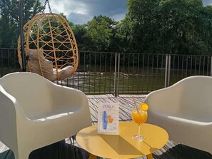 A deck with two white chairs, a hanging chair, a table with a drink and a menu, and a view of a body of water with trees in the background. - Hôtel Mercure Villeneuve sur Lot Moulin de Madame (Villeneuve-sur-Lot)