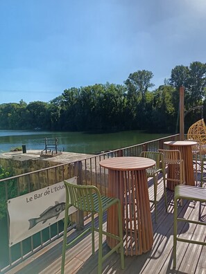 An outdoor dining area with tables and chairs overlooking a body of water, a sign reading 'Le Bar de L'Eclus', and a wooden deck. - Hôtel Mercure Villeneuve sur Lot Moulin de Madame (Villeneuve-sur-Lot)