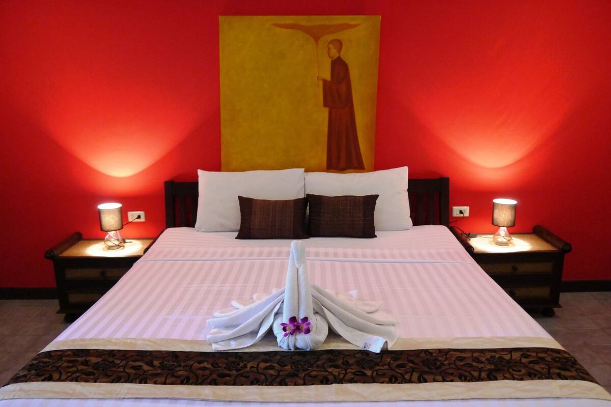 A hotel room with a large bed, two bedside lamps, a red wall, and a religious painting.