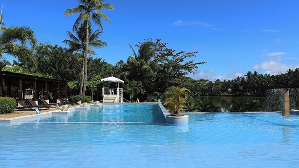 Una piscina con suelo de baldosas, una zona de estar cubierta con sillones de descanso y un pequeño pabellón.