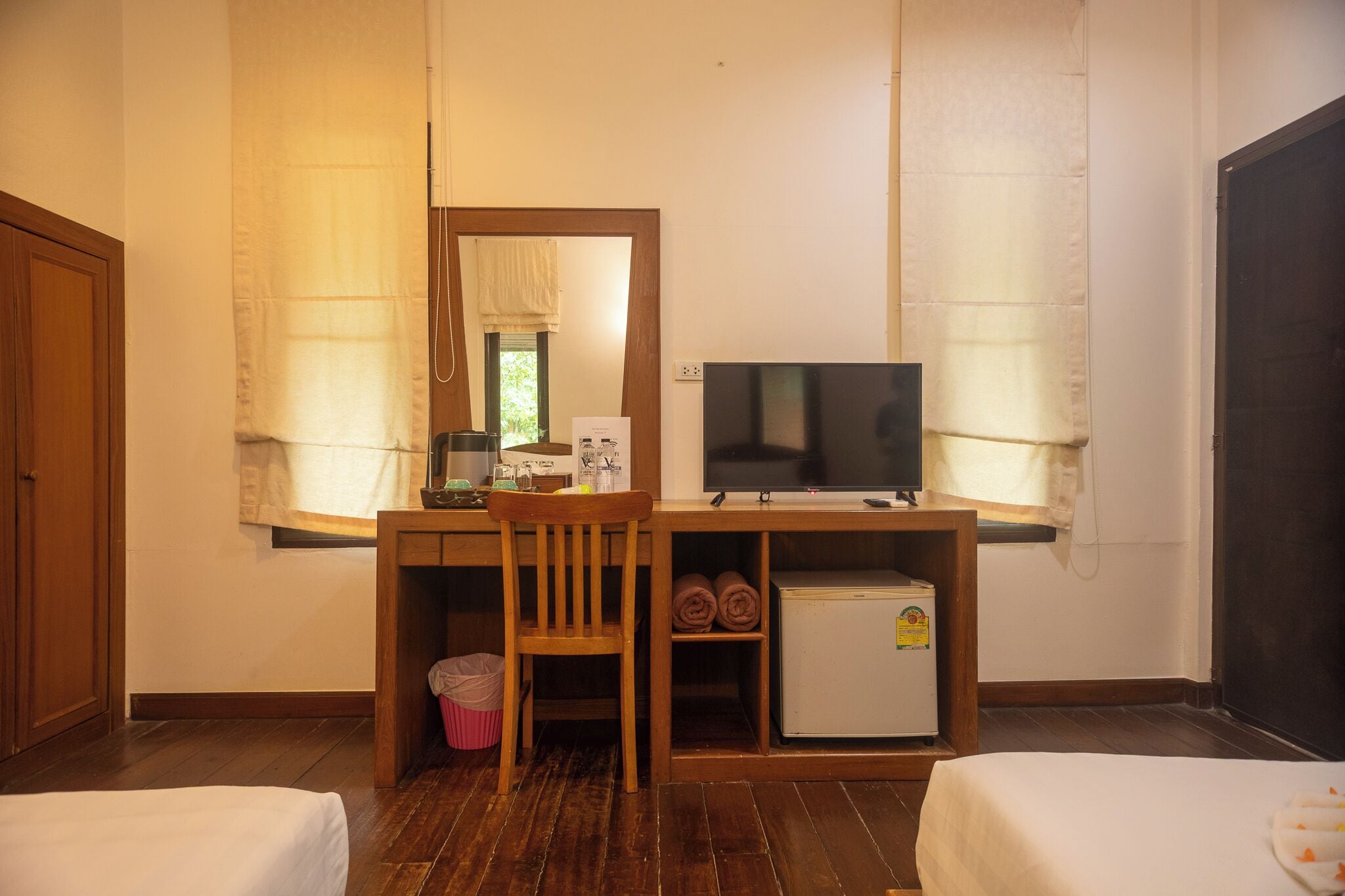 A room with a wooden desk, a television, a small refrigerator, a chair, and two beds.