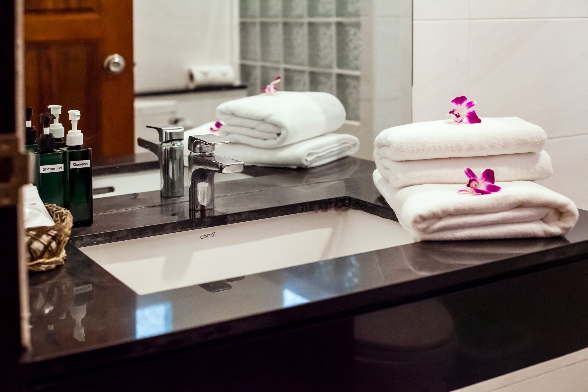 A bathroom with a black countertop, a sink, and a mirror. There are two bottles of shower gel and shampoo, a folded white towel, and a small basket.