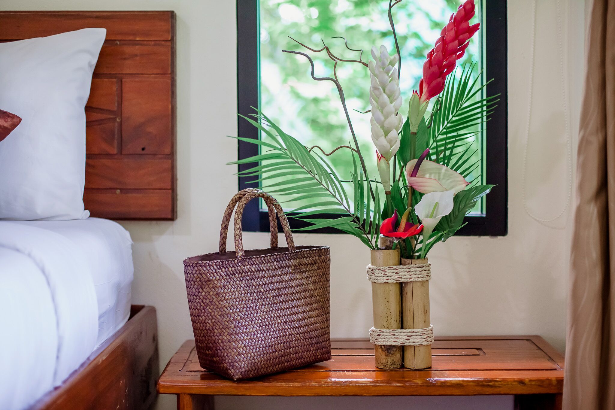 A bed with white bedding, a woven basket, and a vase with flowers on a wooden nightstand.