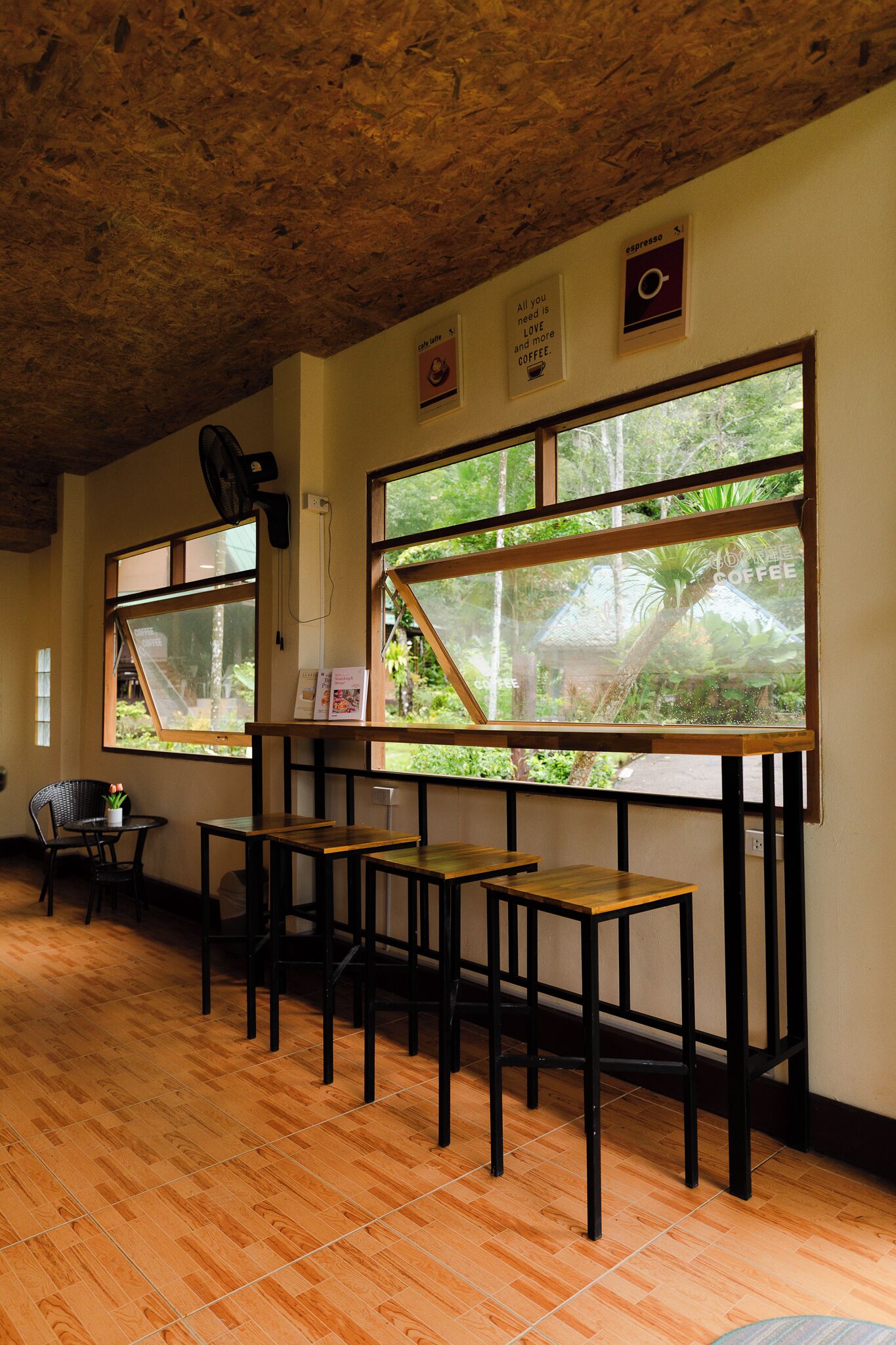 Habitación con pisos y techo de madera, y ventanas amplias con vistas a zonas verdes. Hay mesas y sillas dispuestas para sentarse.
