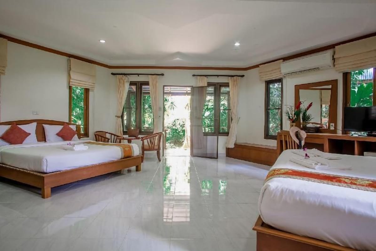 A hotel room with two beds, a desk, a TV, and a view of greenery.