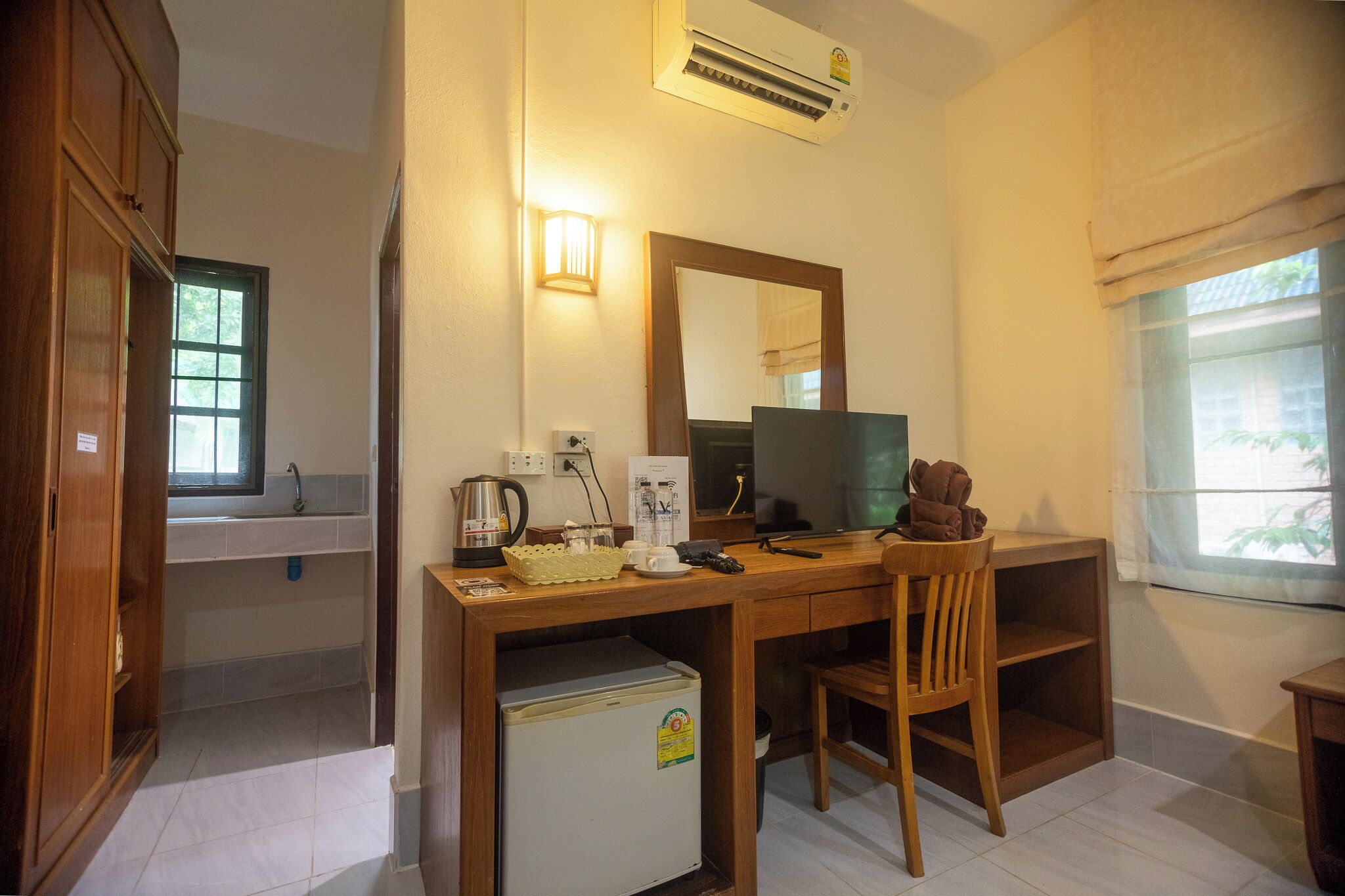 A compact room with a wooden desk, a computer, a chair, a small refrigerator, a window with curtains, and a sink with a mirror.