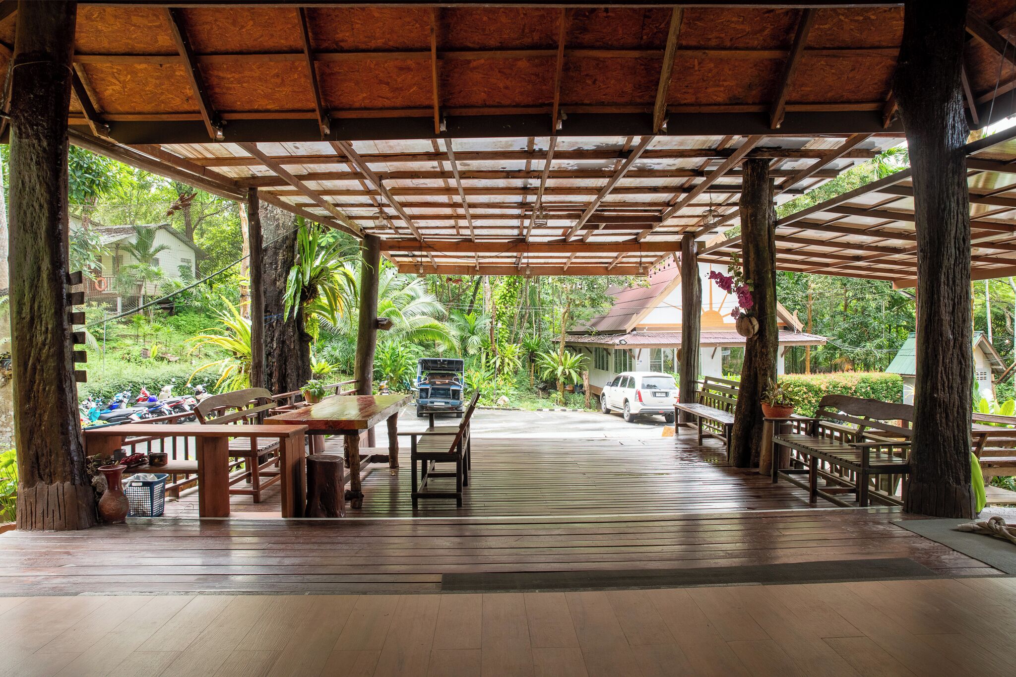 An open-air pavilion with wooden benches and tables, surrounded by lush greenery and a view of a house in the background.