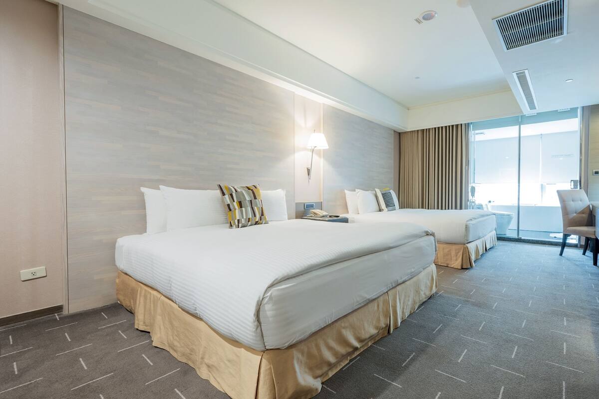 A hotel room with two beds, a desk, a chair, and a large window.