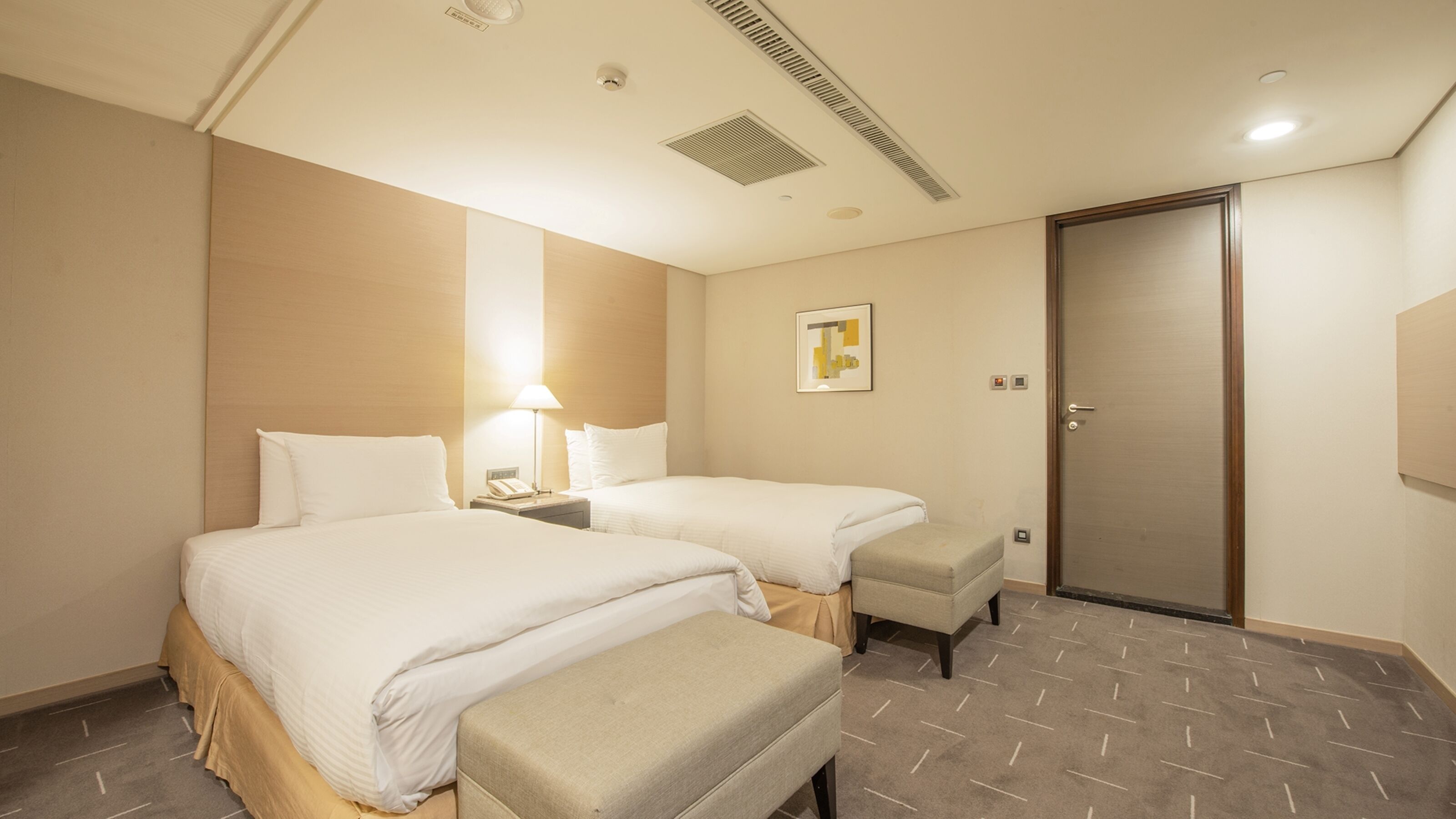 A hotel room with two beds, a bedside table with a lamp, a small bench, a door, and a wall-mounted artwork.