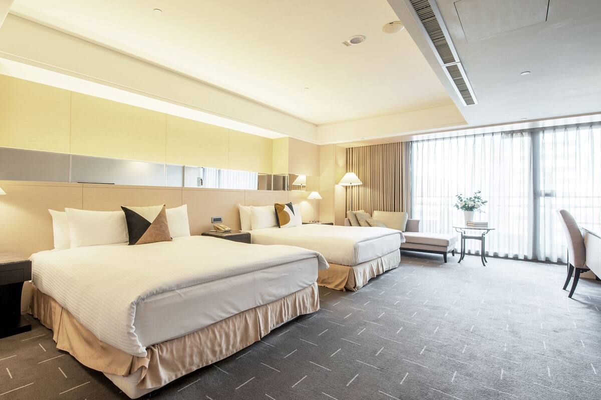A hotel room with two beds, a desk, a chair, a small table with a plant, and large windows with curtains.