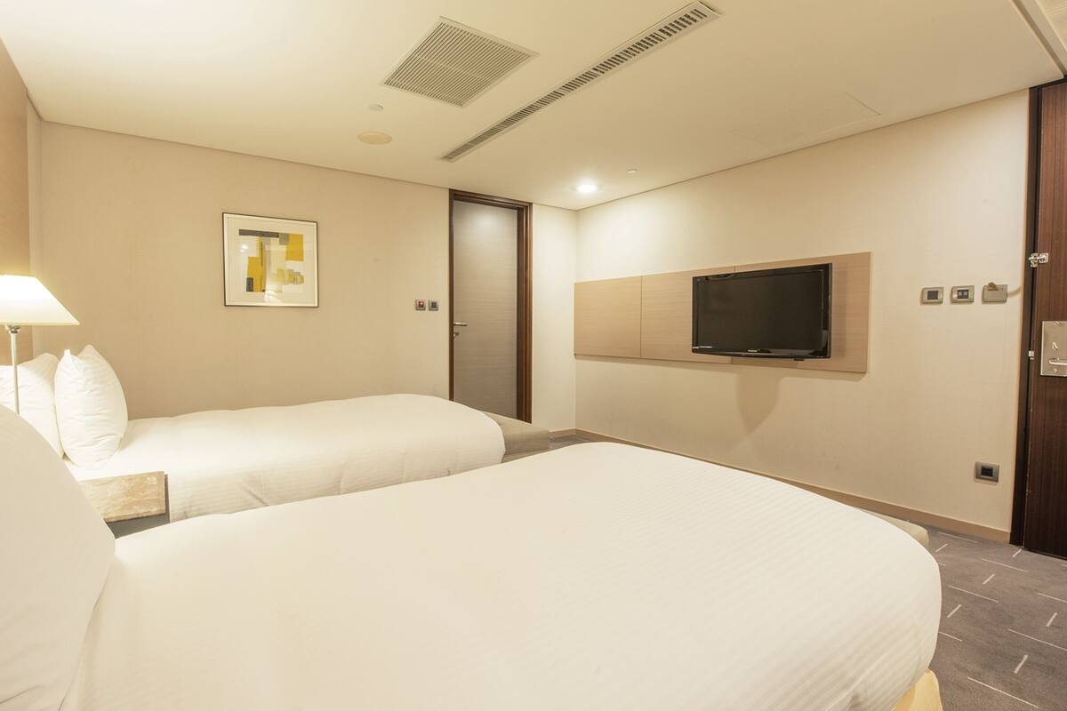 A hotel room with two beds, a TV mounted on the wall, a door, and a ceiling vent.