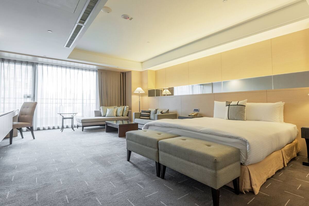 A modern hotel room with a large bed, a desk, a sofa, and a chair.