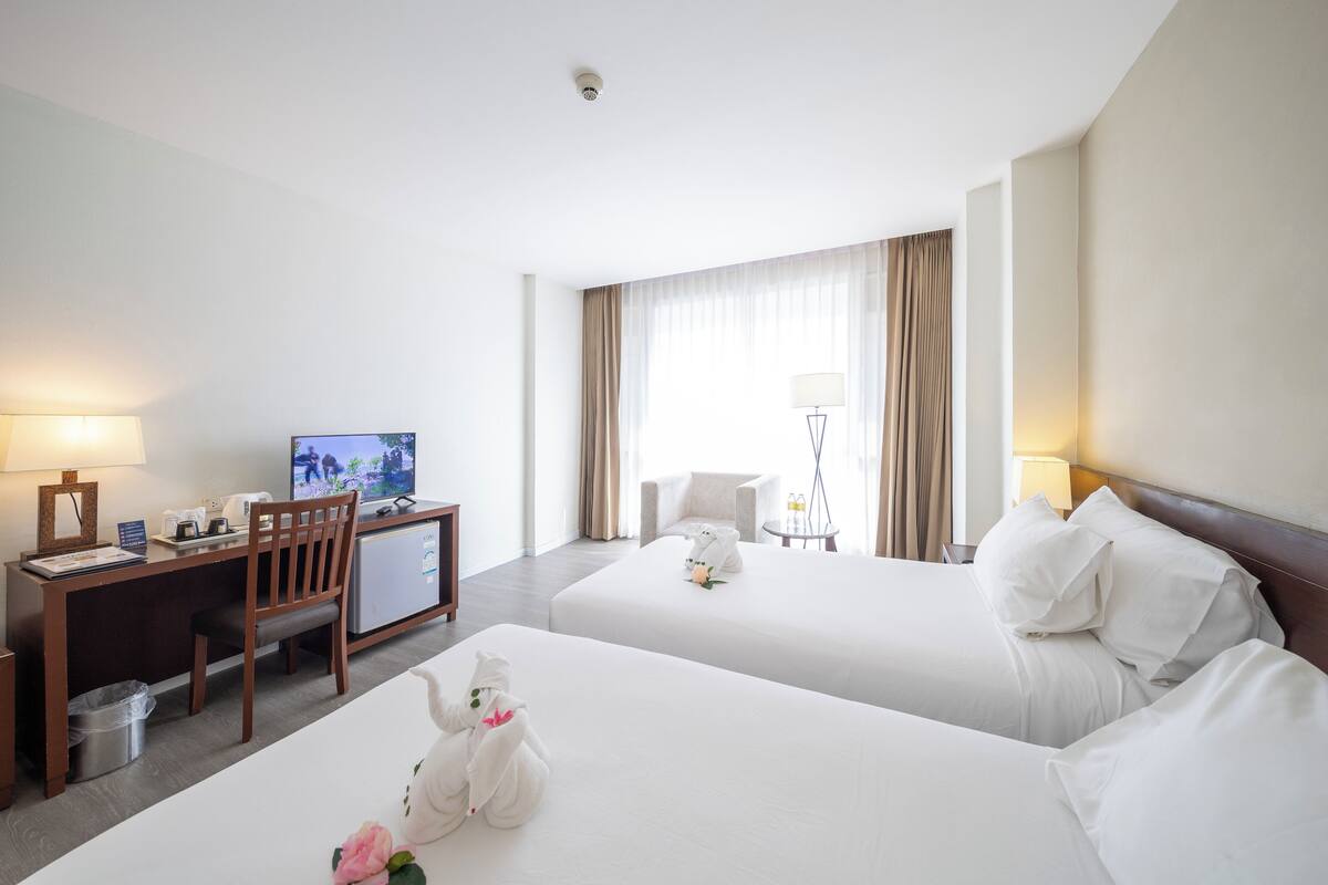 A hotel room with two beds, a desk with a television, a chair, and a small table with a lamp.
