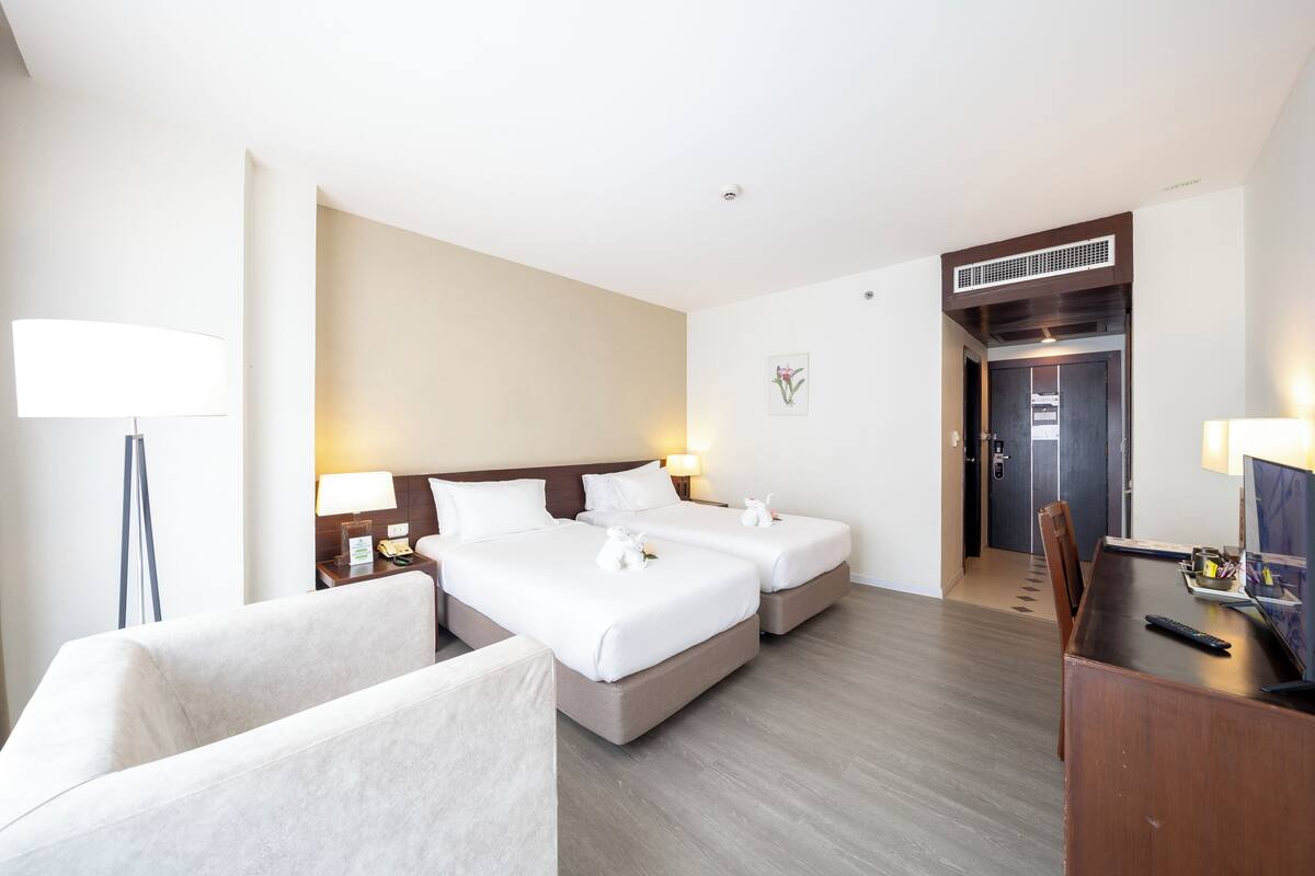A hotel room with two beds, a desk, a chair, a TV, and a lamp.