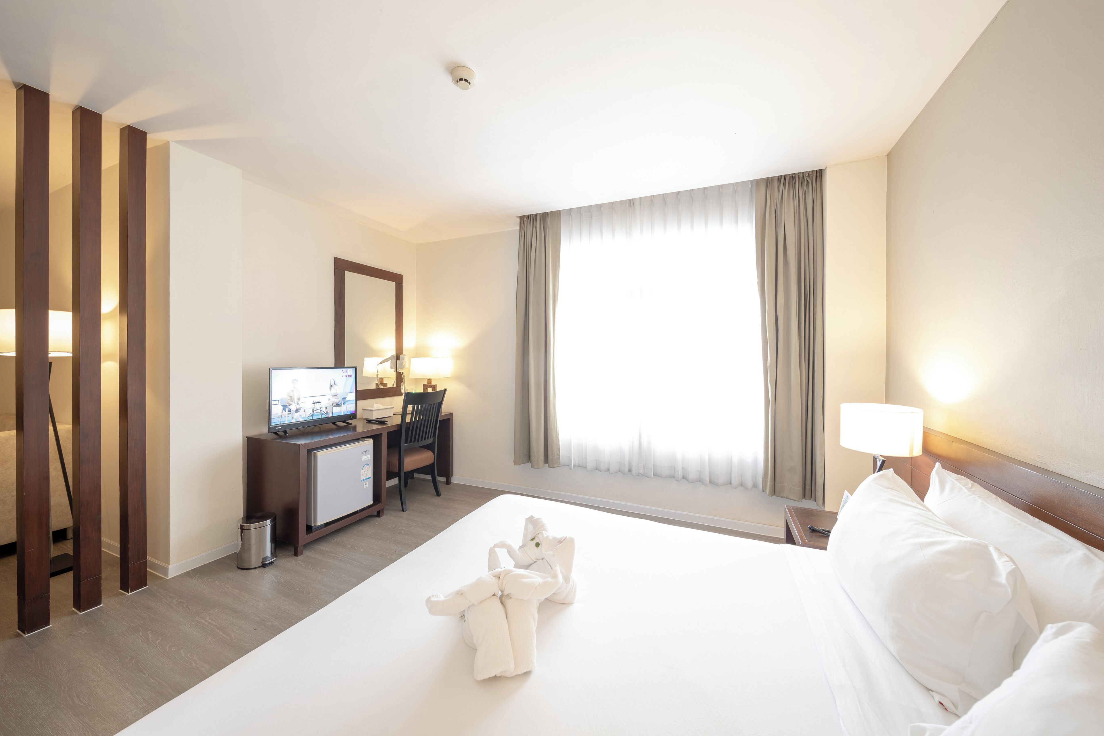 A hotel room with a bed, a desk with a computer, a TV, and a window with curtains.