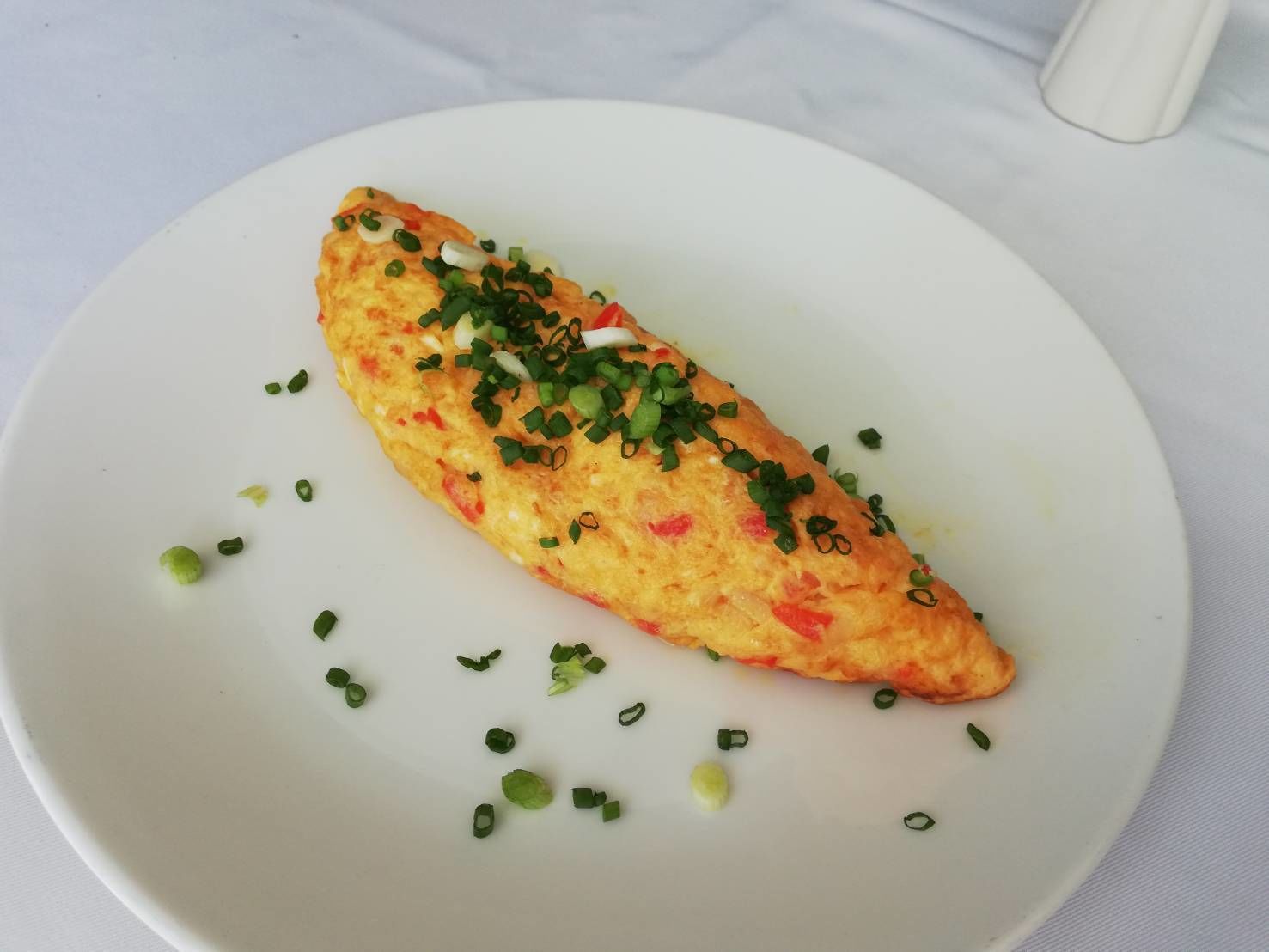 A single omelette on a white plate with chopped green onions and red peppers.
