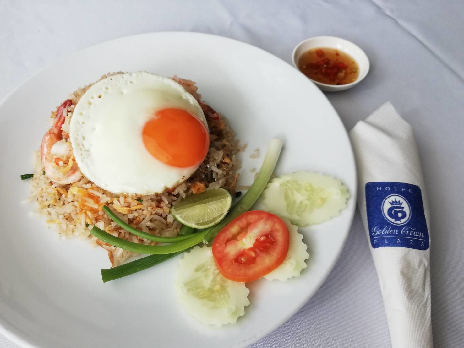 Un plato de arroz frito con camarones, un huevo frito y verduras, acompañado de un recipiente pequeño con salsa y una servilleta con el logotipo de un hotel.